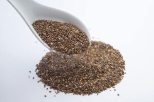 A close-up shot of chia seeds pouring from a white ceramic spoon onto a white surface.